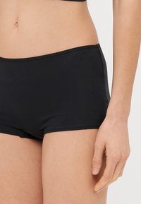 Černé bavlněné hipster kalhotky s hladkým povrchem, elastickým pasem a přiléhavým střihem, částečně zobrazená ruka na boku.