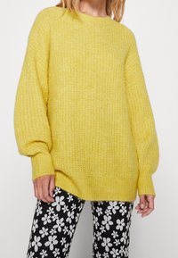 Osoba oblečená v volném žlutém pleteném svetru s balónovými rukávy a černých kalhotách s bílým květinovým vzorem.