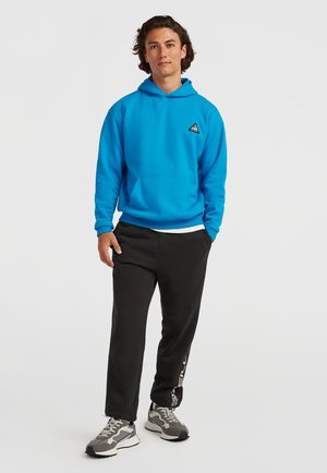 Blue hooded sweatshirt with front pocket, paired with black joggers featuring white lettering. Grey sneakers with a patterned design complete the outfit.