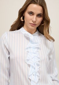 Blusa bianca con righe blu, caratterizzata da un davanti arricciato, chiusura con bottoni e tessuto morbido. Maniche lunghe e scollatura alta.