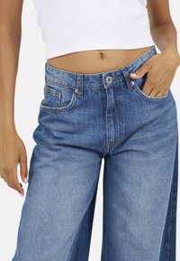Jeans in denim blu con gamba larga e vita alta. Caratteristiche includono cinque tasche, chiusura frontale a bottone e cuciture a vista.