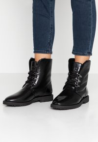 Botas negras hasta el tobillo con un acabado brillante, parte superior texturizada, cierre de cordones en el frente y una suela de goma duradera. Forro de pelaje visible en el borde superior.
