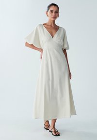 Maxi-jurk met V-hals en korte mouwen in lichtbeige stof, met een geplooide taille en een vloeiende silhouet. Gecombineerd met zwarte sandalen met bandjes.