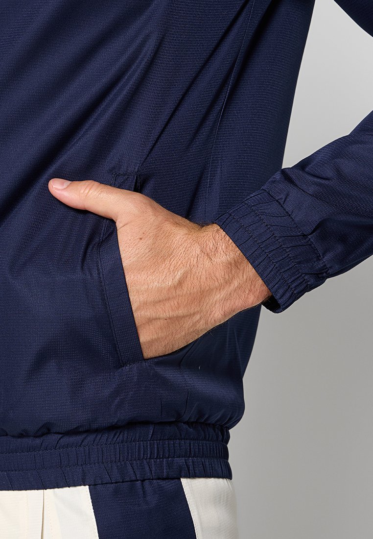 Giacca a vento navy con una texture liscia, dotata di tasca laterale e polsini elasticizzati. Presenta una vestibilità comoda e un tessuto leggero.