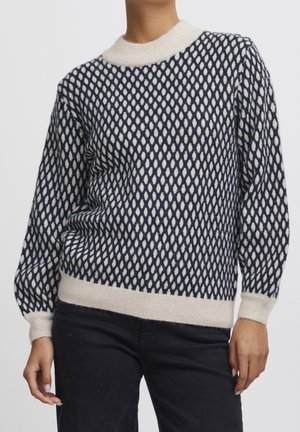 Femme portant un pull en maille à motifs bleu marine et crème avec poignets, ourlet et col côtelés, assorti à un pantalon noir, sur fond uni.