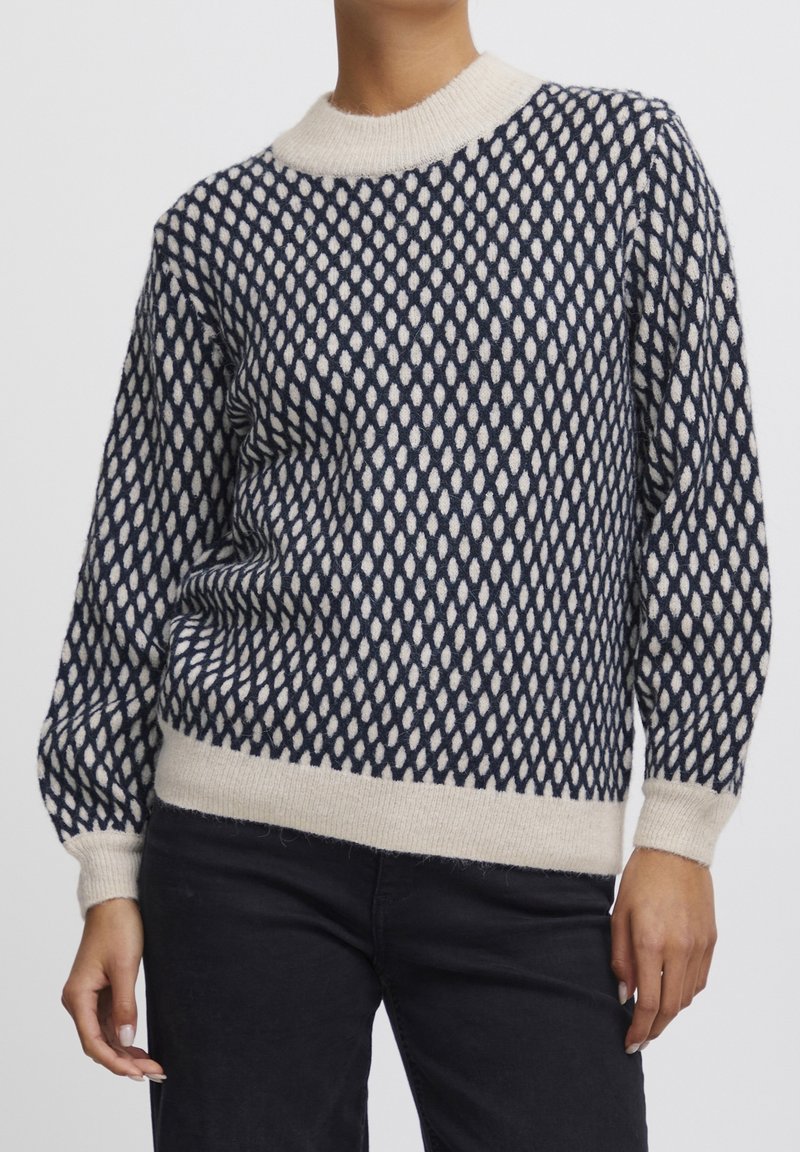 Femme portant un pull en maille à motifs bleu marine et crème avec poignets, ourlet et col côtelés, assorti à un pantalon noir, sur fond uni.