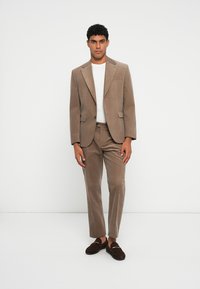 Brązowy garnitur z kordurowego materiału o dopasowanym kroju, z marynarką z klapami i spodniami slim-fit, zestawiony z białą koszulą i ciemnymi loafersami.