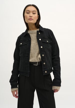 Giubbotto di jeans nero con bottoni argento, tasche frontali e design leggermente accorciato, indossato sopra un maglione di cotone beige.