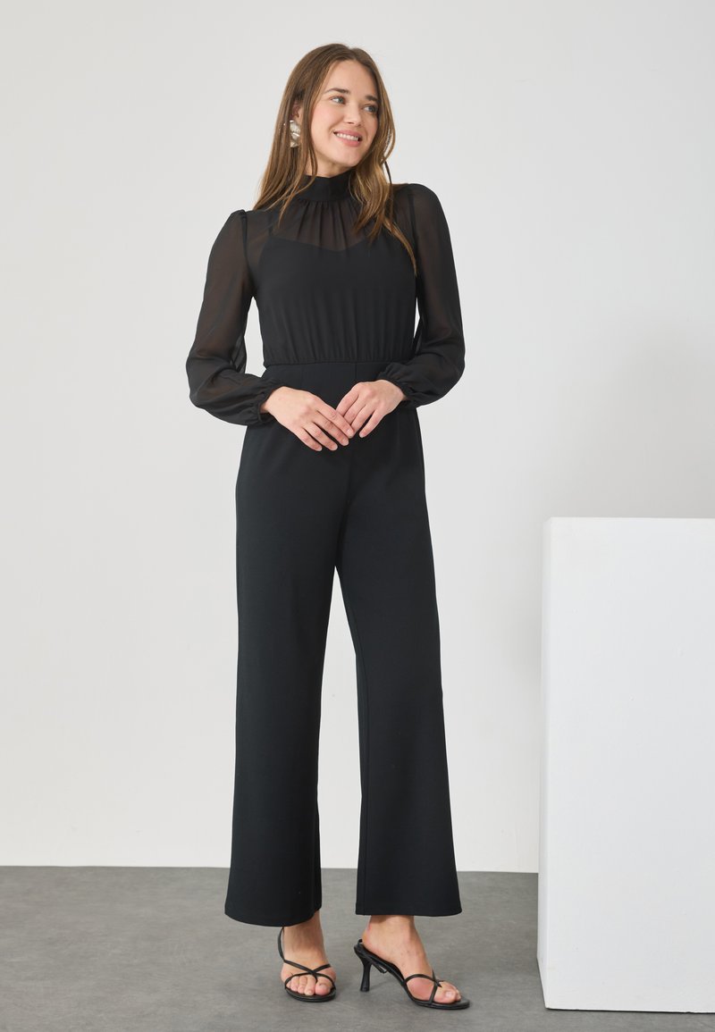 Black jumpsuit with long sheer sleeves, high neckline, and pleated bodice. Relaxed fit legs, worn with black heeled sandals. Simple background.