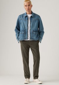 Denim jacket in light blue with two front pockets, button accents, and a slightly cropped cut. Paired with olive green pants and white sneakers.