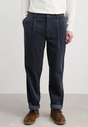 Navy corduroy trousers with a textured, ribbed surface. Features front pleats and rolled cuffs, paired with brown lace-up shoes.