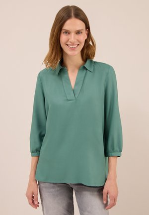 Femme aux cheveux châtain clair mi-long souriant, portant un blouse verte ample à manches 3/4 et un jean gris clair sur fond uni.