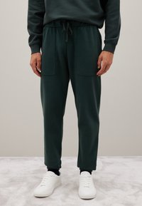 Donkergroene sweatpants met een elastische tailleband, trekkoord en twee voorzakken. Strakke boorden bij de enkels, gedragen met witte sneakers.
