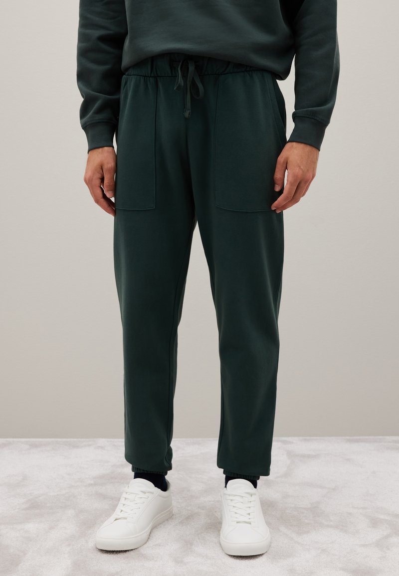 Donkergroene sweatpants met een elastische tailleband, trekkoord en twee voorzakken. Strakke boorden bij de enkels, gedragen met witte sneakers.