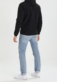 Veste noire légère à capuche avec manches longues, associée à un jean bleu clair et des baskets blanches. Coupe décontractée, sans motifs ni logos apparents.