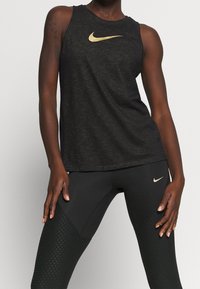 Czarna bezrękawnikowa koszulka na ramiączkach z złotym logo Nike. Noszona z czarnymi legginsami z teksturowanym wzorem diamentowym i detalami akcentującymi.