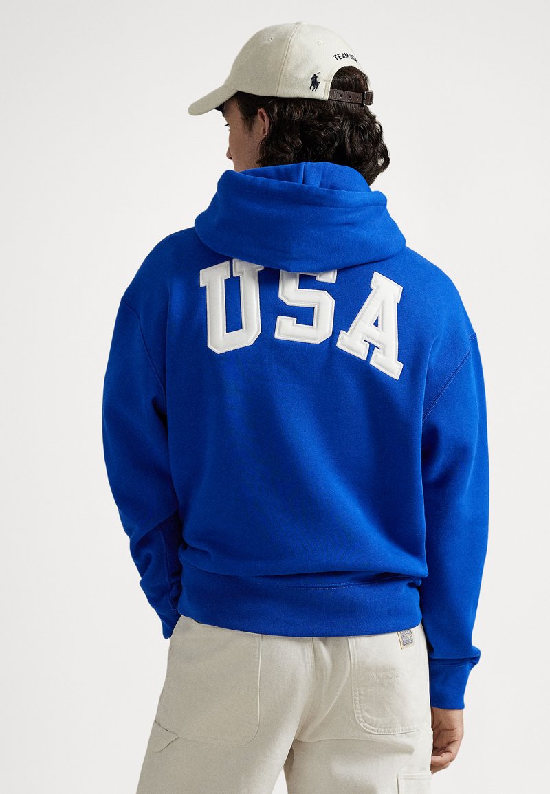 Blå hettegenser med en stor, hvit "USA" sydd på ryggen. Mykt materiale med romslig hette og ribbede mansjetter. Beige caps på hodet.