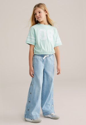 Fată tânără cu păr blond lung, purtând un top scurt verde mentă cu numărul „08” și blugi evazați bleu deschis cu nasturi laterali.