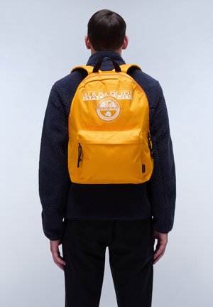 Gelber Rucksack mit einem Logo und einem Aufnäher auf der Rückseite, ausgestattet mit verstellbaren Trägern und einer Fronttasche, kombiniert mit einer marineblauen Fleecejacke.