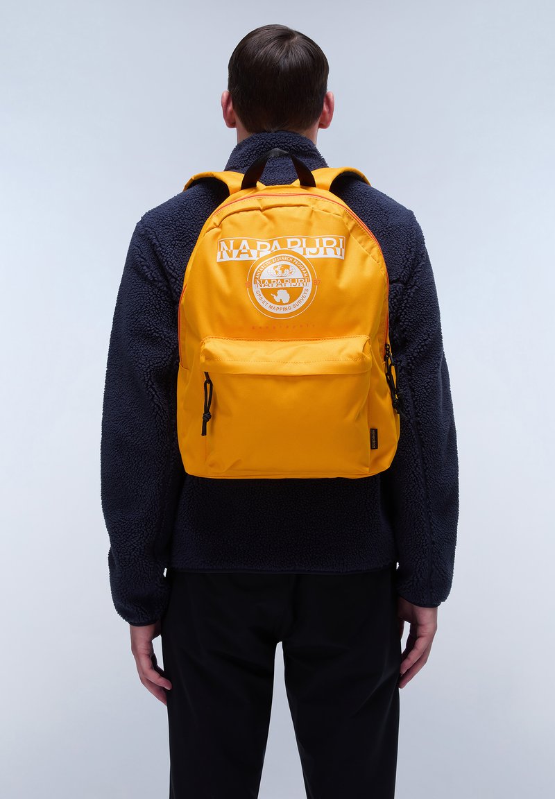 Mochila amarilla con un logo y parche en la parte posterior, con correas ajustables y un bolsillo delantero, combinada con una chaqueta de forro polar azul marino.