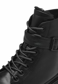 Bottine en cuir noir avec des lacets gris, une sangle latérale et des œillets en métal. Dispose d'une doublure intérieure douce pour le confort et d'un design à bout rond.