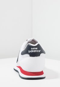 White New Balance sneaker with black heel tab, red midsole stripe, and black outsole, shown from rear view on white surface.