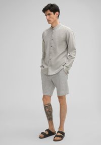 Helles graues Hemd mit langen Ärmeln und Knöpfen, kombiniert mit gestreiften Shorts und schwarzen Riemchensandalen. Hat einen Stehkragen und eine strukturierte Oberfläche.