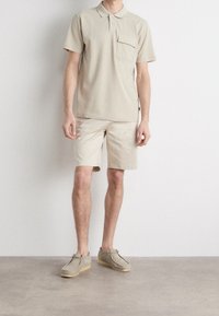 Polo en beige à manches courtes avec une poche, associé à un short beige et des chaussures gris clair, présentant une texture et un design décontracté.