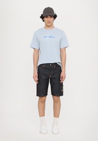 Light blue short-sleeve t-shirt with graphic print, dark denim shorts with cargo pockets, and white sneakers, accessorized with a grey cap.