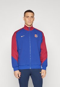 Blå och maroon zip-up jacka med hög krage, Nike-logotyp och FC Barcelona-emblem. Slät tyg, långa ärmar och ribbade mudd.