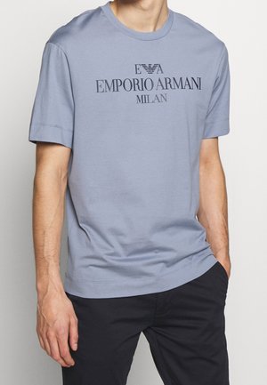 Jasnoniebieska bawełniana koszulka z krótkimi rękawami. Z przodu znajduje się czarny nadrukowany tekst logo "EMPORIO ARMANI MILAN". Gładka textura.