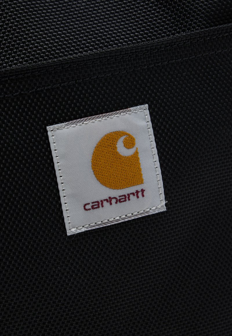Etichetta di tessuto bianco con logo Carhartt arancione e testo rosso cucita su sfondo di tessuto nero strutturato.