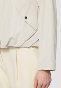 Veste coupe-vent beige clair avec une fermeture éclair, détails de poches, texture lisse et manches longues, associée à un pantalon de couleur claire.