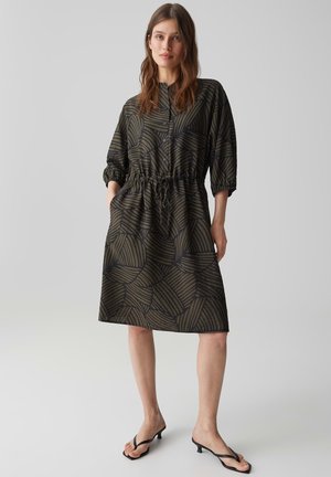 Femme portant une robe longue aux genoux à manches longues avec des motifs géométriques vert foncé et noir, debout les mains dans les poches et des sandales noires à petit talon.