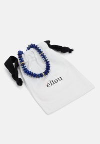 Pulseira de contas azuis com pedras redondas e brilhantes e detalhes em ouro, colocada sobre um saco de tecido branco com a marca "éliou."