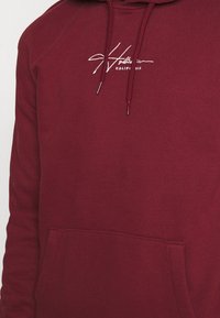 Sudadera burdeos de tela suave con un bolsillo frontal tipo canguro, cordones y el logotipo bordado de "Hollister California" en blanco.