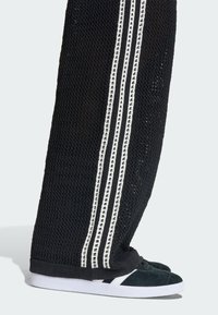 Čierne sieťované nohavice s bielymi zvislými pásikmi a textúrovaným pletením; doplnené tmavozelenými teniskami s bielou podrážkou a detailmi.