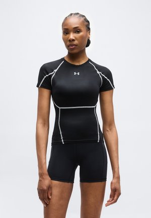 Athletische Frau steht, nach vorne blickend, trägt ein eng anliegendes schwarzes Kurzarmshirt und passende schwarze Shorts mit weißen Nähten.