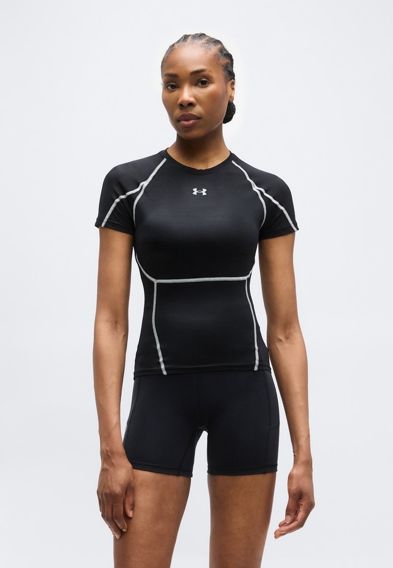 Αθλητική γυναίκα που στέκεται στραμμένη προς τα εμπρός, φορώντας μαύρο εφαρμοστό κοντομάνικο τοπ και μαύρο σορτς με λευκά ραφές.