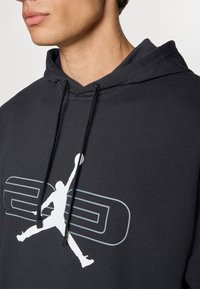 Sort hoodie med kænguru-lomme, der har et hvidt silhuetlogo og grå tekst, lavet af et struktureret stof med justerbare snore.