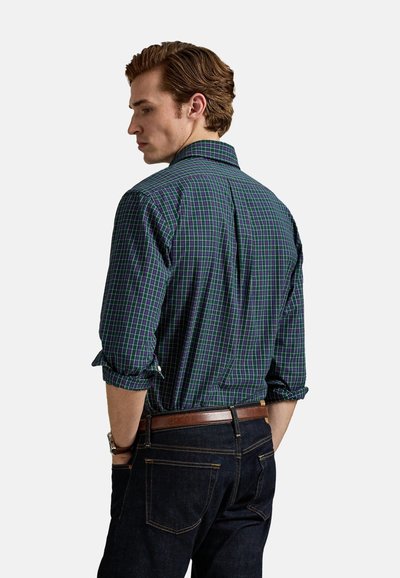 Camisa de manga larga a cuadros con patrón en tonos verde oscuro, azul y morado, mangas remangadas y detalle en la espalda, combinada con jeans vaqueros oscuros.