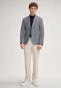 Blazer a quadri grigio e bianco, chiusura a un bottone, dolcevita nera sotto, abbinato a pantaloni beige chiaro e sneaker bianche.