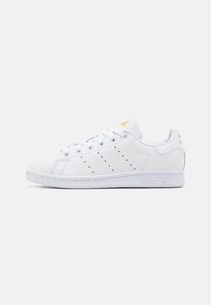 Valkoinen Adidas Stan Smith -tennari rei'itetyillä raitoja, valkoisilla nauhoilla ja kultaisilla logokoristeilla kannassa ja kielen päällä, sivuprofiilissa.