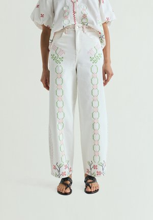 Witte broek met verticale pastelroze en groene bloemenborduursel, gecombineerd met zwarte sandalen met studs en een wit geborduurd bloesje.