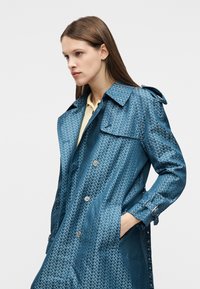 Νεαρή γυναίκα με ίσια καστανά μαλλιά φοράει ένα μπλε παλτό με σχέδια πάνω από ένα ανοιχτό κίτρινο τοπ, με το ένα χέρι στην τσέπη του παλτού, κοιτάζοντας αριστερά.