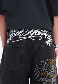 Schwarze Denim-Shorts mit weißem, gesticktem Text auf dem Bund und bunten Strass-Applikationen eines Tigerdesigns an der Seite.