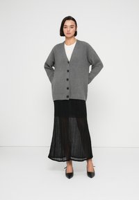 Γκρι πλεκτή ζακέτα με μακριά μανίκια, με V λαιμόκοψη και πέντε σκούρα κουμπιά. Συνδυασμένη με μαύρη maxi φούστα με πιέτες και μαύρες γόβες.