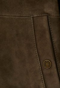 Material din suede maro cu o textură moale, cu cusături proeminente și un buton circular din alamă cu numele mărcii gravat.