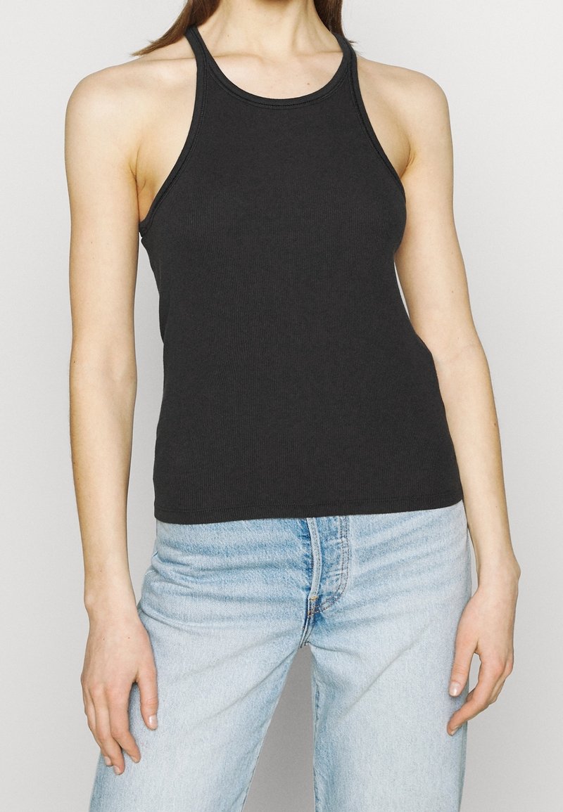 Frau in einem schwarzen ärmellosen Tanktop, kombiniert mit hellblauen hoch taillierten Jeans, vor einem schlichten hellen Hintergrund stehend.