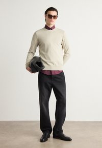 Svetlo bež pleteni pulover, maroon srajca na gumb, črni denim hlače, črne čevlje in sončna očala. Moški drži črno torbo. Minimalistična ozadje.
